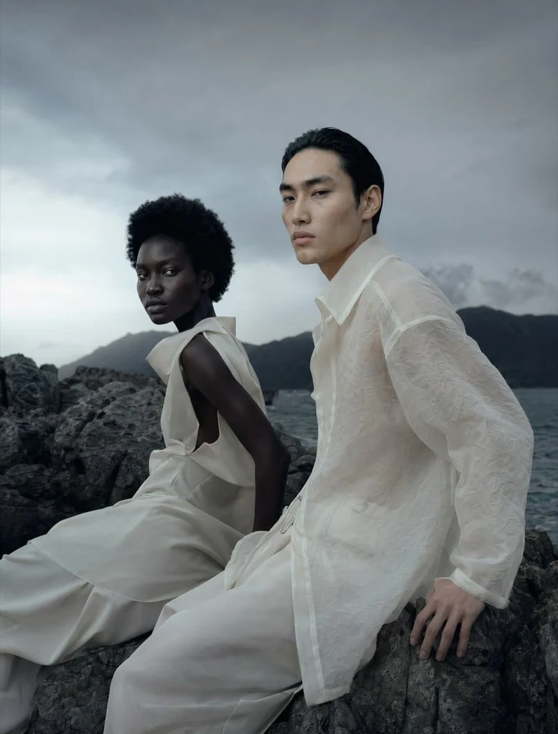 AI-generated on-model image: two models in sheer white layers on a coastal rock
